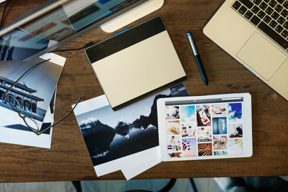 A creative workspace with a laptop, tablet, stylus, and printed photos scattered across a wooden desk.